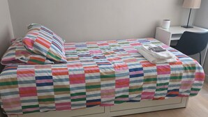 Un letto singolo con copriletto a righe, un cuscino e un asciugamano piegato. A destra, una scrivania con una sedia e una lampada.