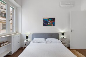 Ein Schlafzimmer mit einem Bett, zwei Nachttischen mit Lampen, einem Fenster, einer Tür und einem farbenfrohen, abstrakten Gemälde an der Wand.