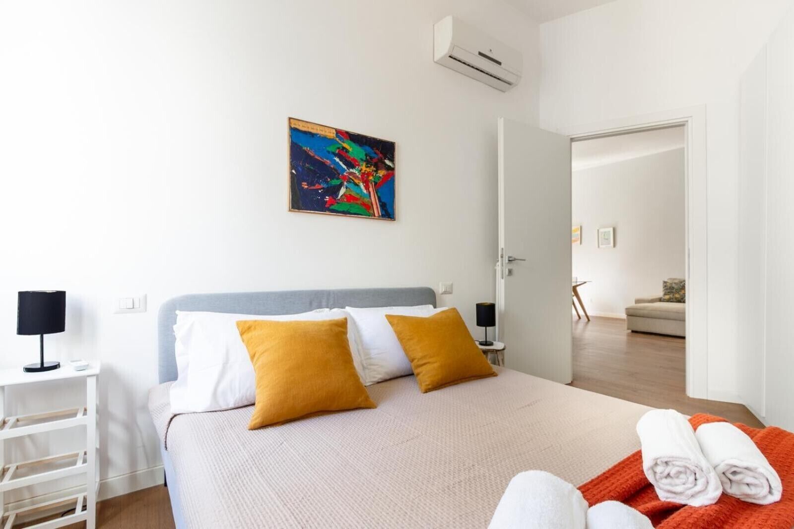 Une chambre d’hôtel avec un lit, une table de chevet, une lampe, un tableau et une porte donnant sur une autre pièce.