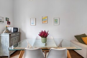 Ein Essbereich mit einem Glastisch, weißen Stühlen, einer Blumenvase und Kunstwerken an der Wand.