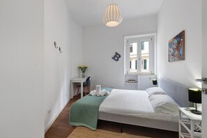 Une chambre à coucher comprenant un lit, une table de chevet, une lampe, une fenêtre et un petit bureau avec une chaise.
