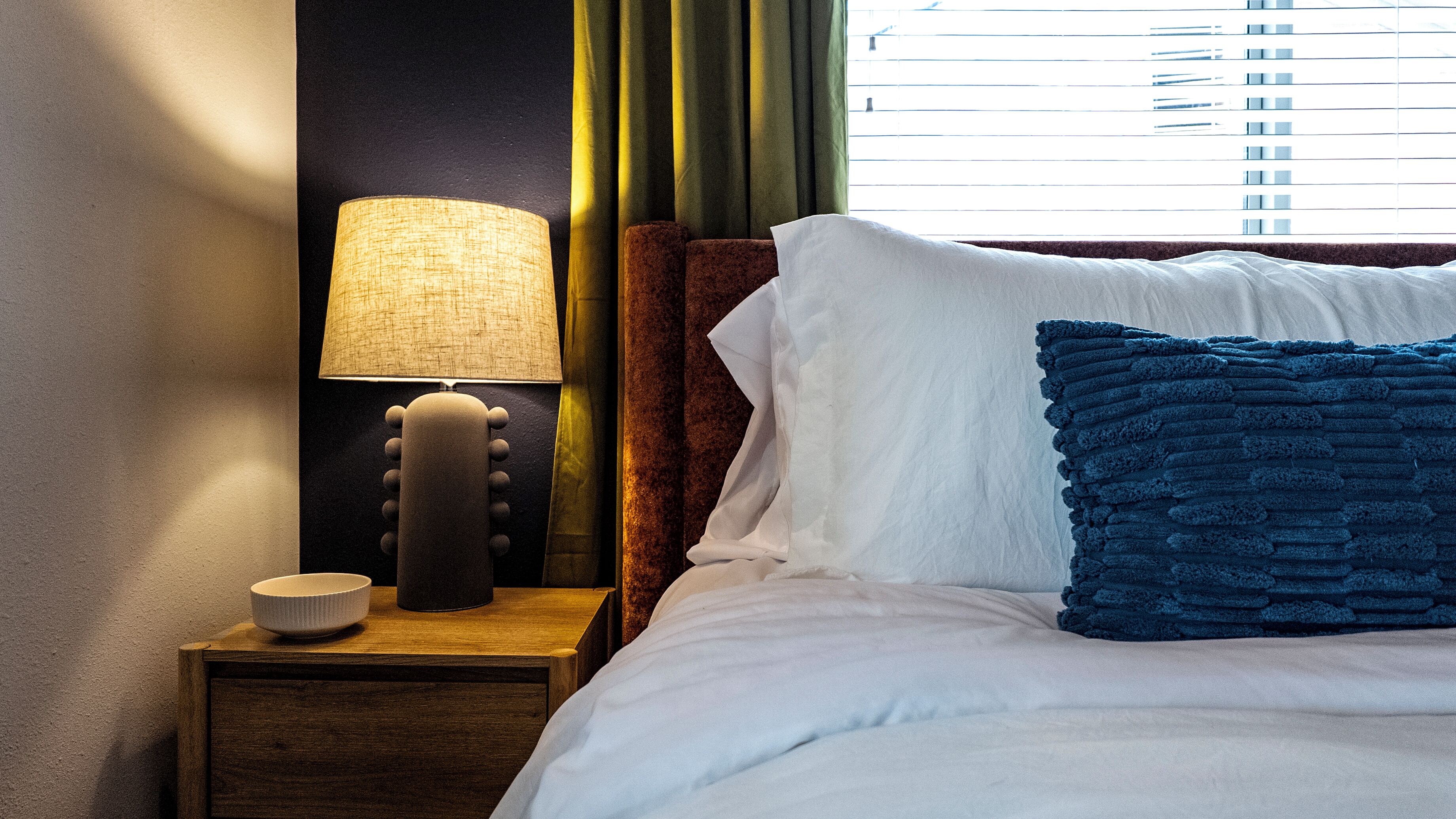 Un lit bien fait, avec des oreillers blancs et bleus, une lampe de chevet et une petite table sur laquelle se trouve un bol.