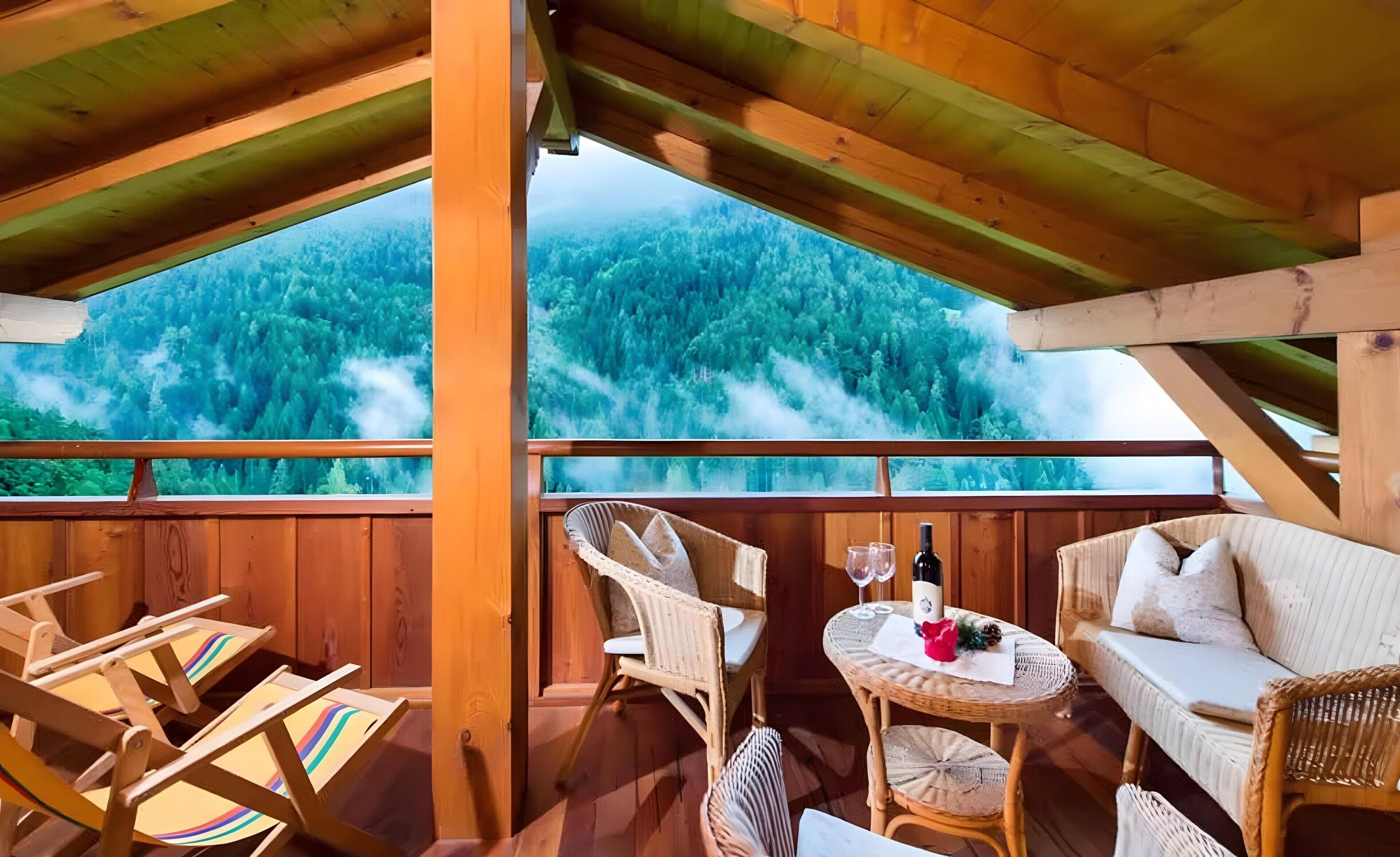 Eine Holzterrasse mit Blick auf einen Wald, Korbmöbel und ein Tisch mit einer Flasche und Gläsern.