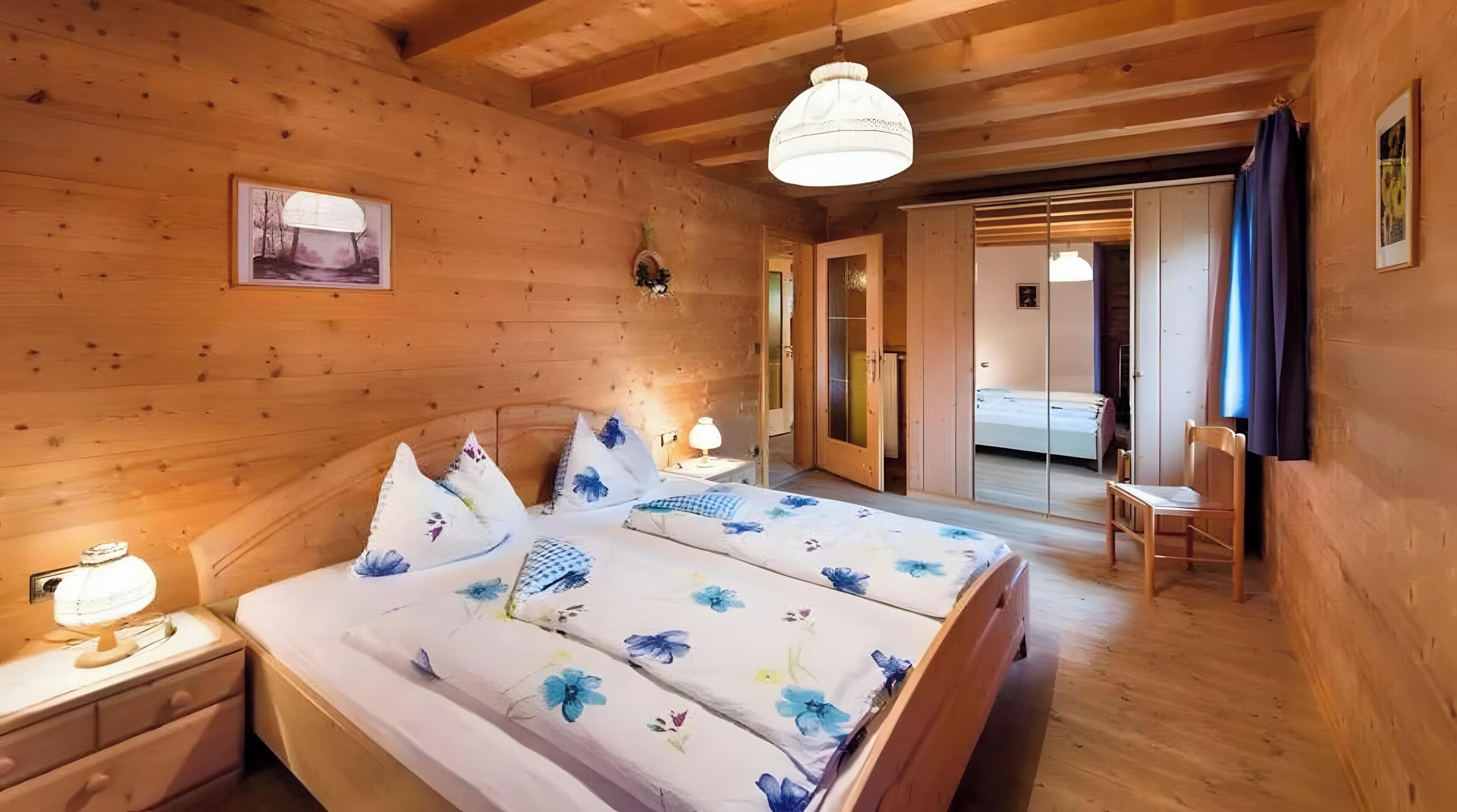 Ein Zimmer mit Holzboden, einem Bett, Nachttischen, einer Hängeleuchte und einer Tür zu einem anderen Raum.