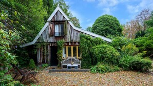 Una casa de madera rústica con techo inclinado, una banca afuera y un jardín con hojas caídas.