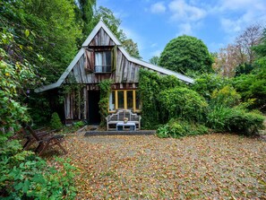 Una casa de madera rústica con techo inclinado, una banca afuera y un jardín con hojas caídas.