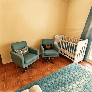 A room with two chairs and a crib, a quilted bed, and a tiled floor.