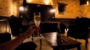 A hand holding a champagne glass in a dimly lit room with stone walls and a fireplace. - 1503 – Exceptional Suite for Two in the Heart of Historic Sarlat-France (Sarlat la Canéda)