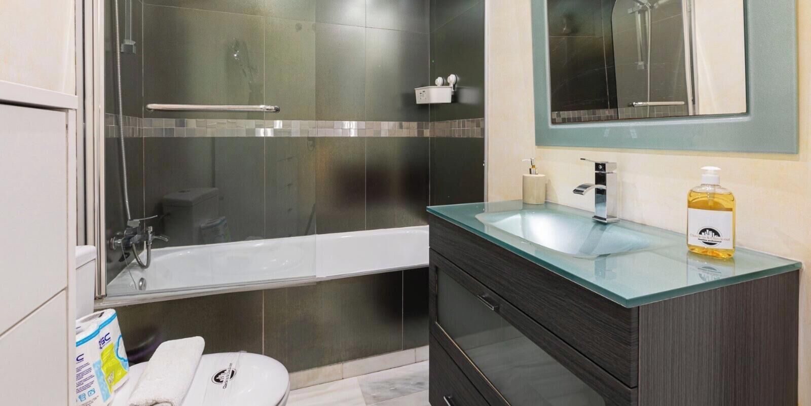 Un baño moderno con ducha de vidrio, una bañera blanca, lavamanos de vidrio y muebles de color oscuro.