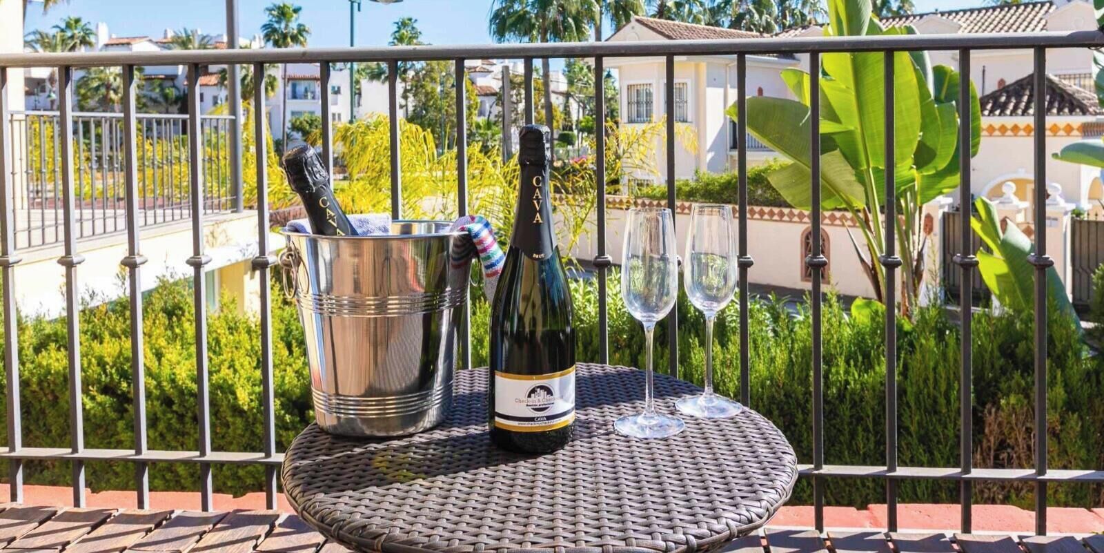 A metal table with a bottle of Cava wine and two glasses on a balcony with a metal railing, surrounded by greenery and buildings.