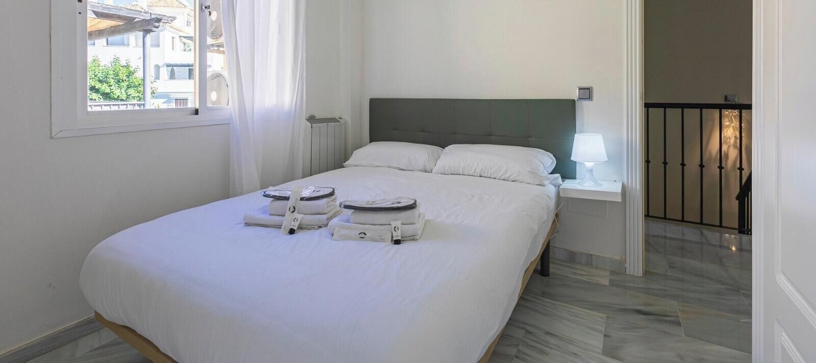 A hotel room with a bed, a nightstand, a lamp, and a view of the outside.