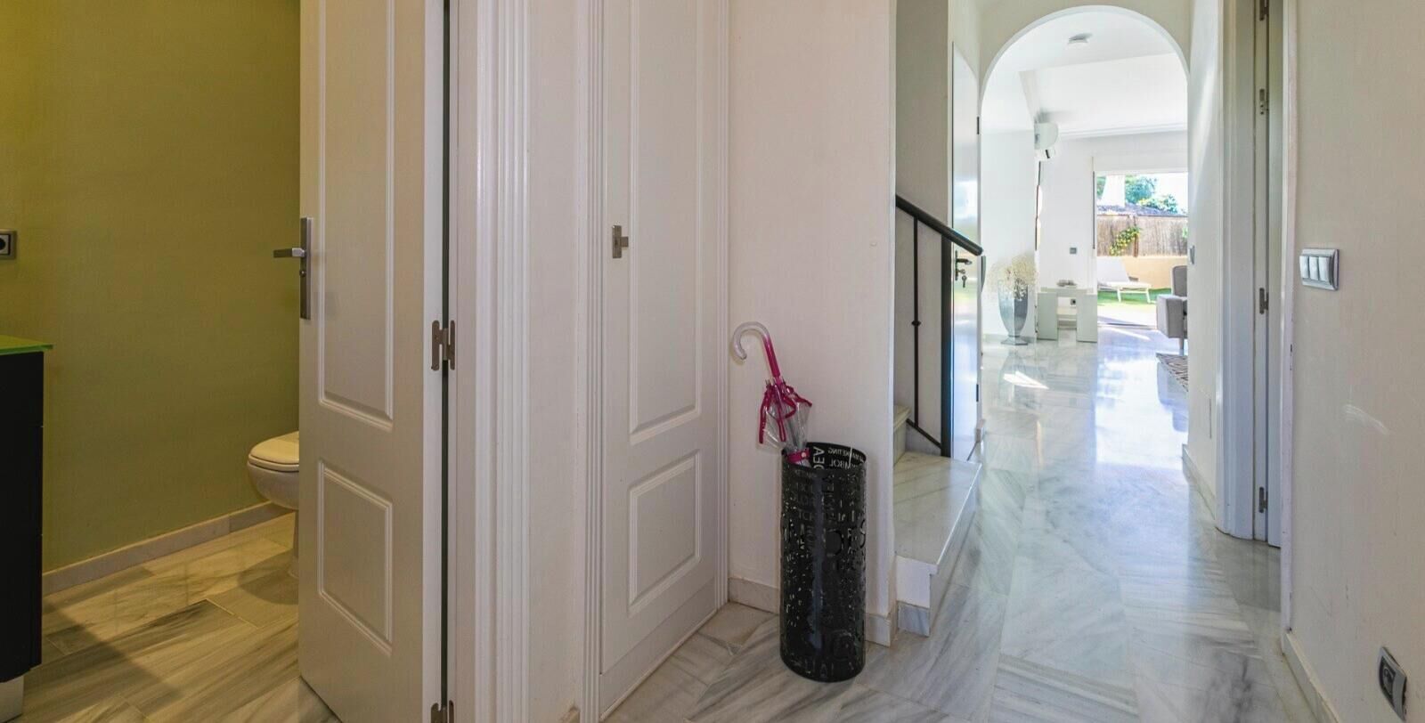 Un pasillo con una puerta blanca, un basurero y vistas a una habitación luminosa con una escalera.