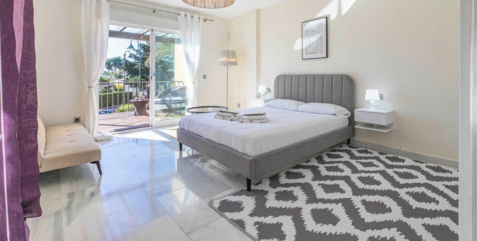 Un dormitorio con cama, mesita de noche, lámpara y una alfombra estampada.