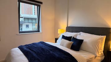 A hotel room with a large bed, a window with a view of buildings, a bedside lamp, and a dark headboard.