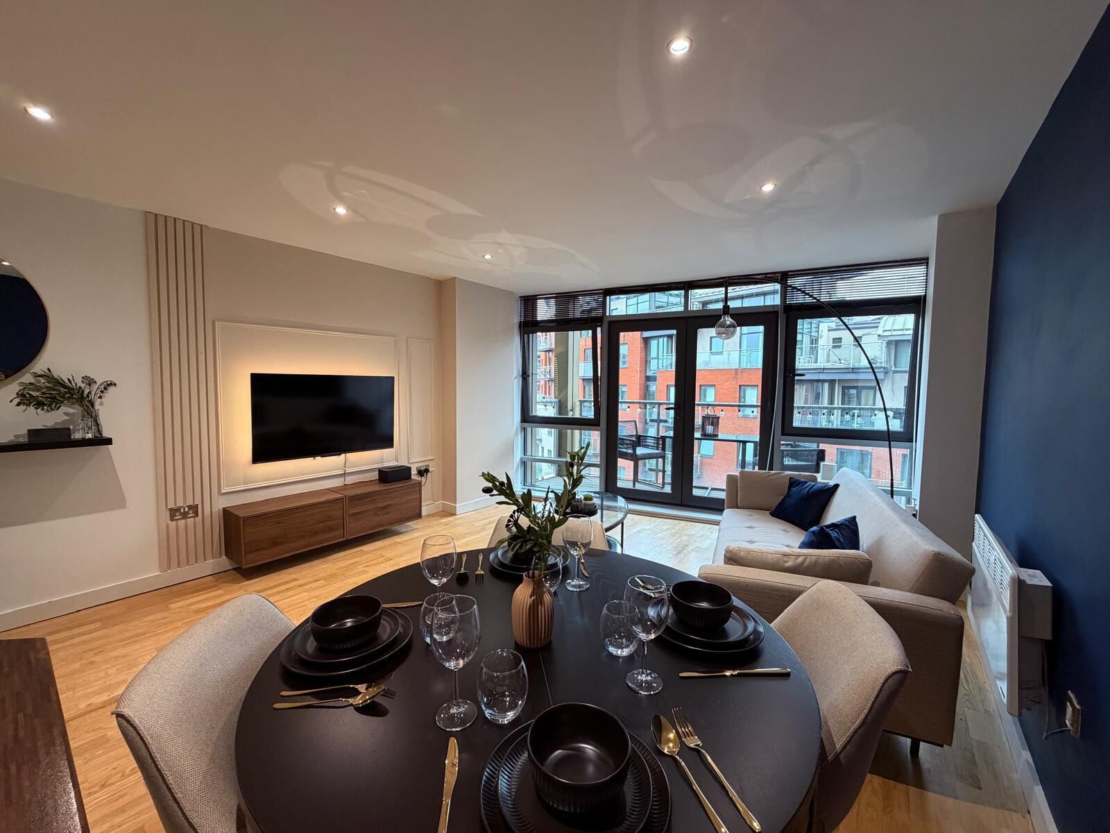 Un espace repas moderne avec une table ronde pour quatre personnes, un téléviseur à écran plat fixé au mur et une grande fenêtre donnant sur la ville.