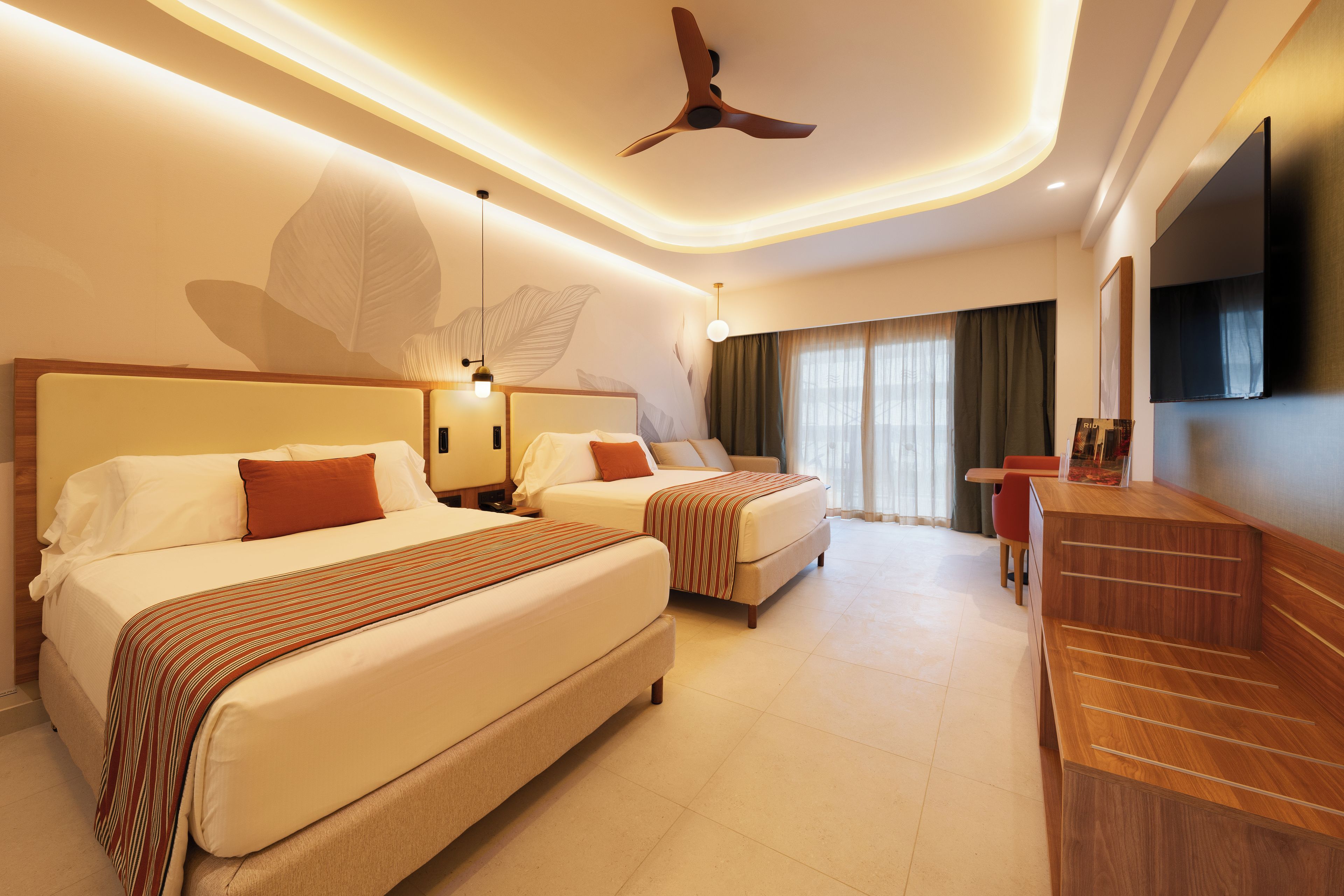 Une chambre d’hôtel avec deux lits, un ventilateur de plafond, une télévision et une grande fenêtre avec des rideaux.