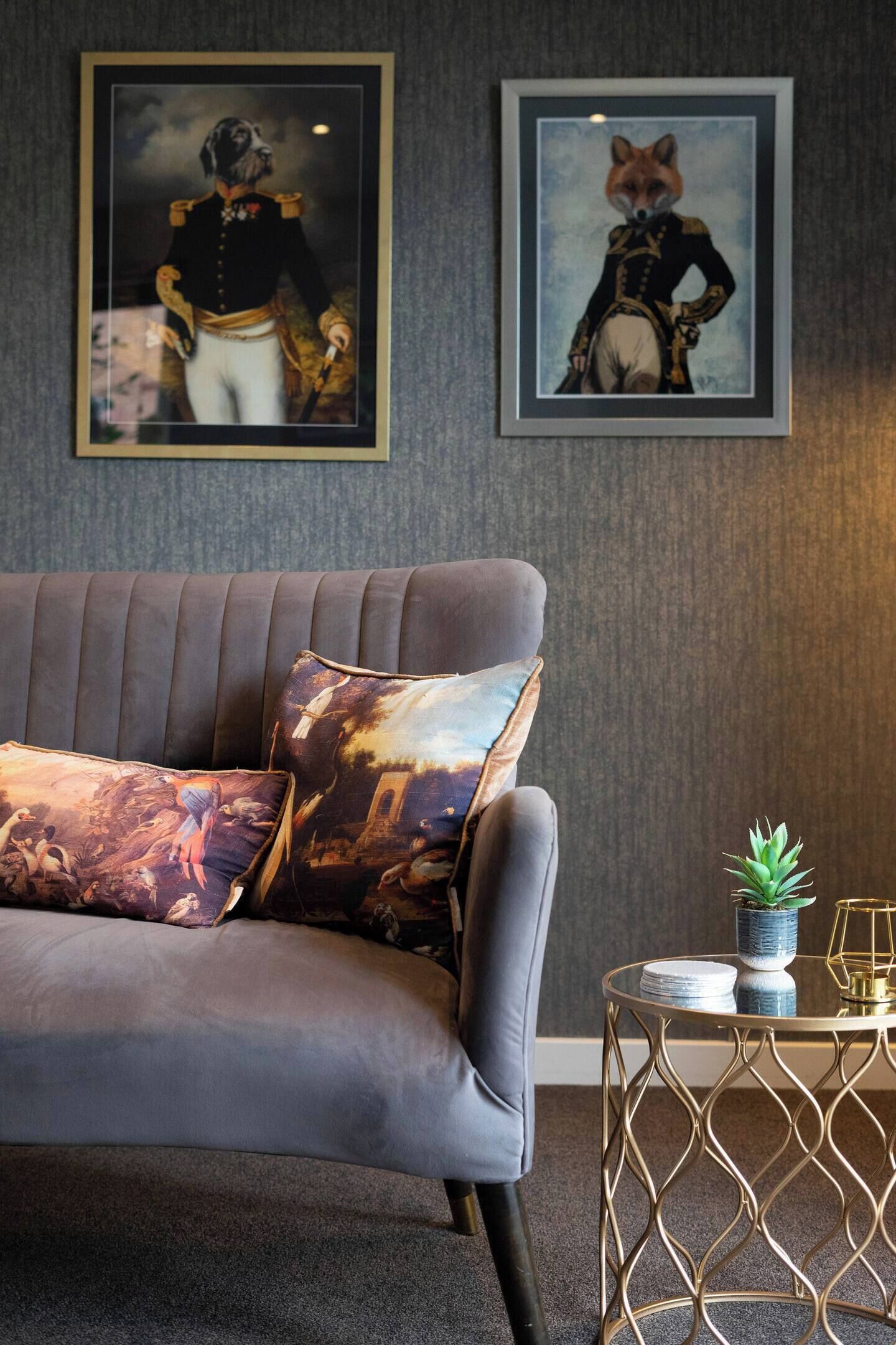 A grey sofa with patterned cushions, a framed artwork of a dog in uniform, and a small table with a plant and a glass object.