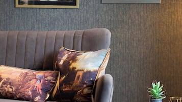 A grey sofa with patterned cushions, a framed artwork of a dog in uniform, and a small table with a plant and a glass object.