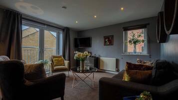 Un salon comprenant un canapé, un fauteuil et une table basse. Un téléviseur est fixé au mur et une fenêtre offre une vue sur un immeuble.