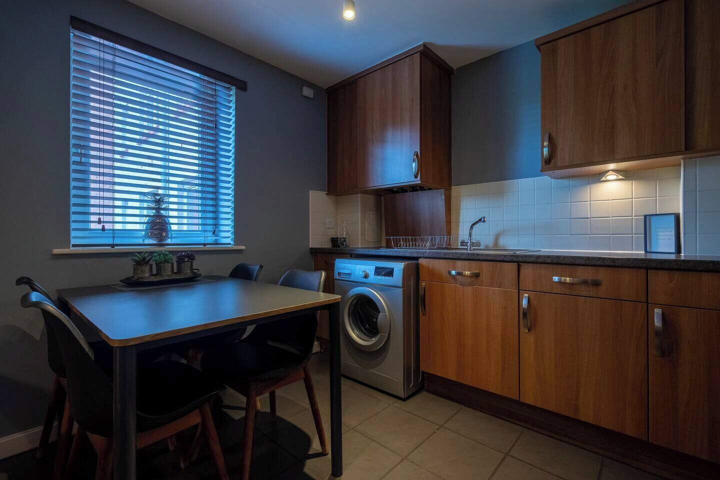 A compact kitchen with a washing machine, wooden cabinets, a dining table with chairs, and a window with blinds.