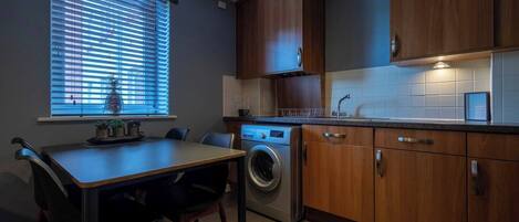 A compact kitchen with a washing machine, wooden cabinets, a dining table with chairs, and a window with blinds.