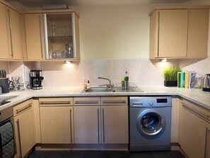 A compact kitchen with wooden cabinets, a built-in dishwasher, a washing machine, and a small sink area. - Our apartment is close to the City Centre and Leith (Edinburgh)