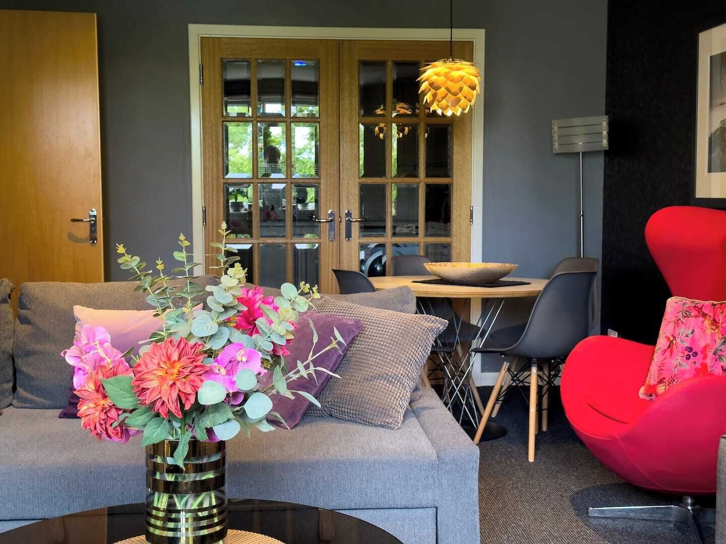 A modern living room with a grey sofa, a red chair, a vase with flowers, and a dining area with a table and chairs.