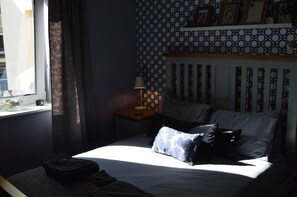 Un dormitorio con una cama, una mesita de noche con lámpara, una ventana con cortinas y un papel tapiz estampado.