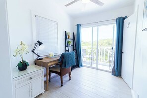 Ein Zimmer mit einem Holztisch, einem Stuhl, einem weißen Schminktisch, einem Fenster mit Jalousien, einem Deckenventilator und Blick ins Grüne.