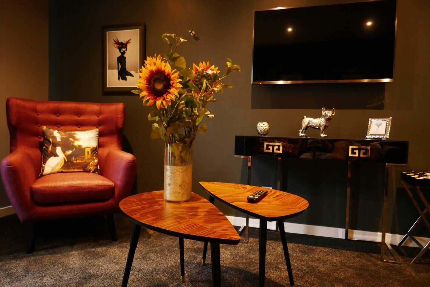 Une pièce avec un fauteuil rouge, une table basse en bois, un vase rempli de tournesols, un téléviseur à écran plat et un trophée posé sur une console.