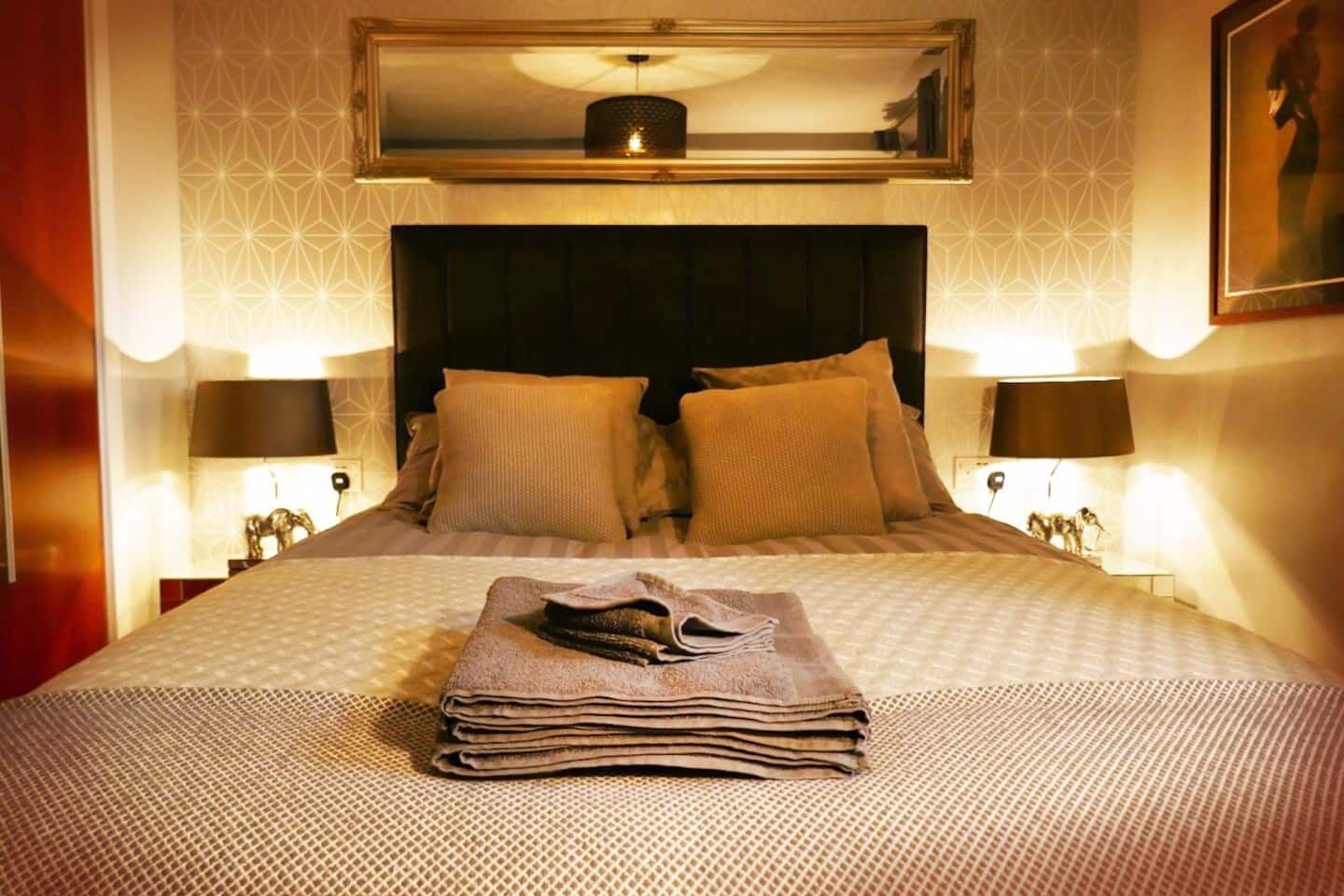 A neatly made bed with a folded towel, two bedside lamps, and a framed mirror above the bed.
