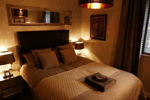 A hotel room with a large bed, a headboard, two bedside lamps, a nightstand, a mirror, and a framed picture on the wall. - Luxury boutique apartment decorated to a high standard (Edinburgh)