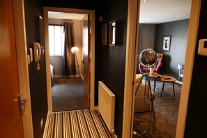 Ein Hotelgang mit Holztür, Streifen Teppich, Stativ mit Spiegel und einem Raum mit Bett und Vorhängen im Hintergrund.