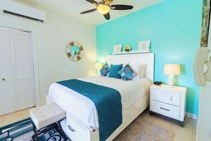 Ein Schlafzimmer mit einem Bett, Nachttischen, einem Nachtschränkchen, einem Deckenventilator, einer Wanduhr und einer Tür mit Klappläden.