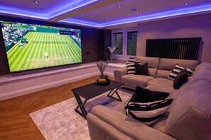 Ein Wohnzimmer mit einem großen Fernseher, der ein Tennismatch zeigt, einem beigefarbenen Sofa, einem kleinen Couchtisch und einem Fenster mit Jalousien.