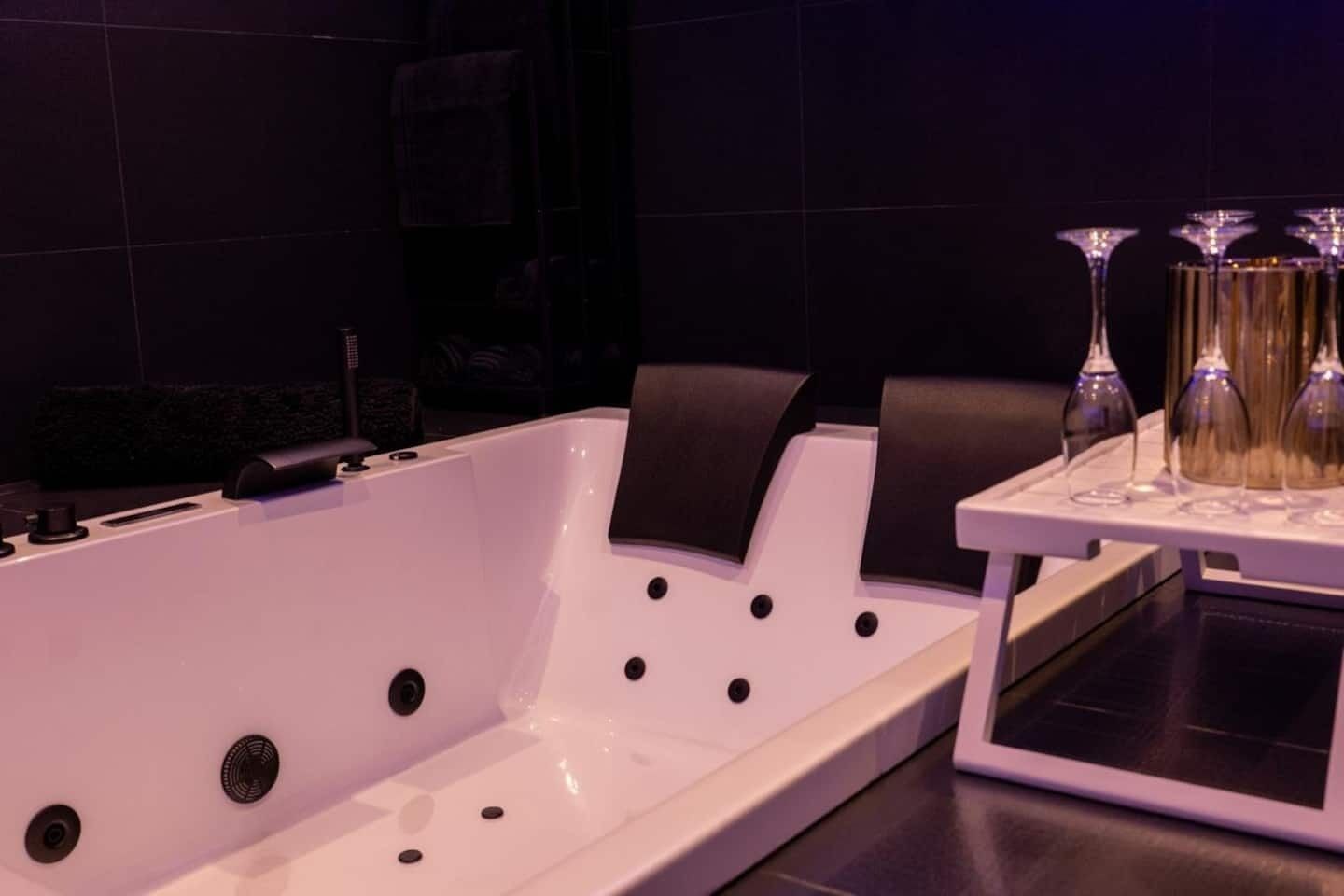 A modern bathroom with a white bathtub, black chairs, and a tray with wine glasses.
