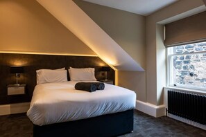A modern bedroom with a large bed, a sloped ceiling, a window with a stone wall view, and a radiator. - Sophisticated Urban Living| Sleek 4-Bedroom Home (Edinburgh)