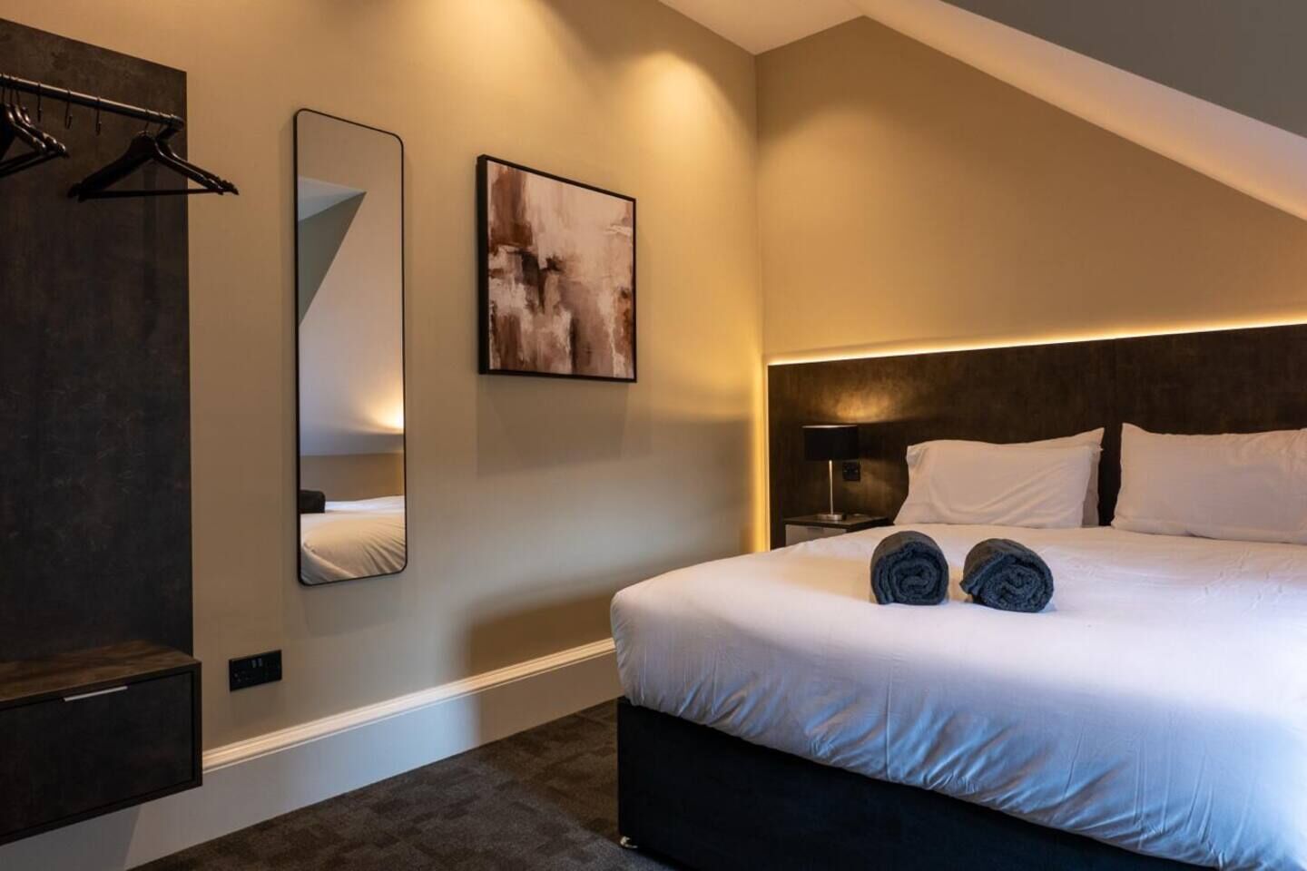 A modern hotel room with a large bed, a bedside table with a lamp, a wall-mounted mirror, and a painting.