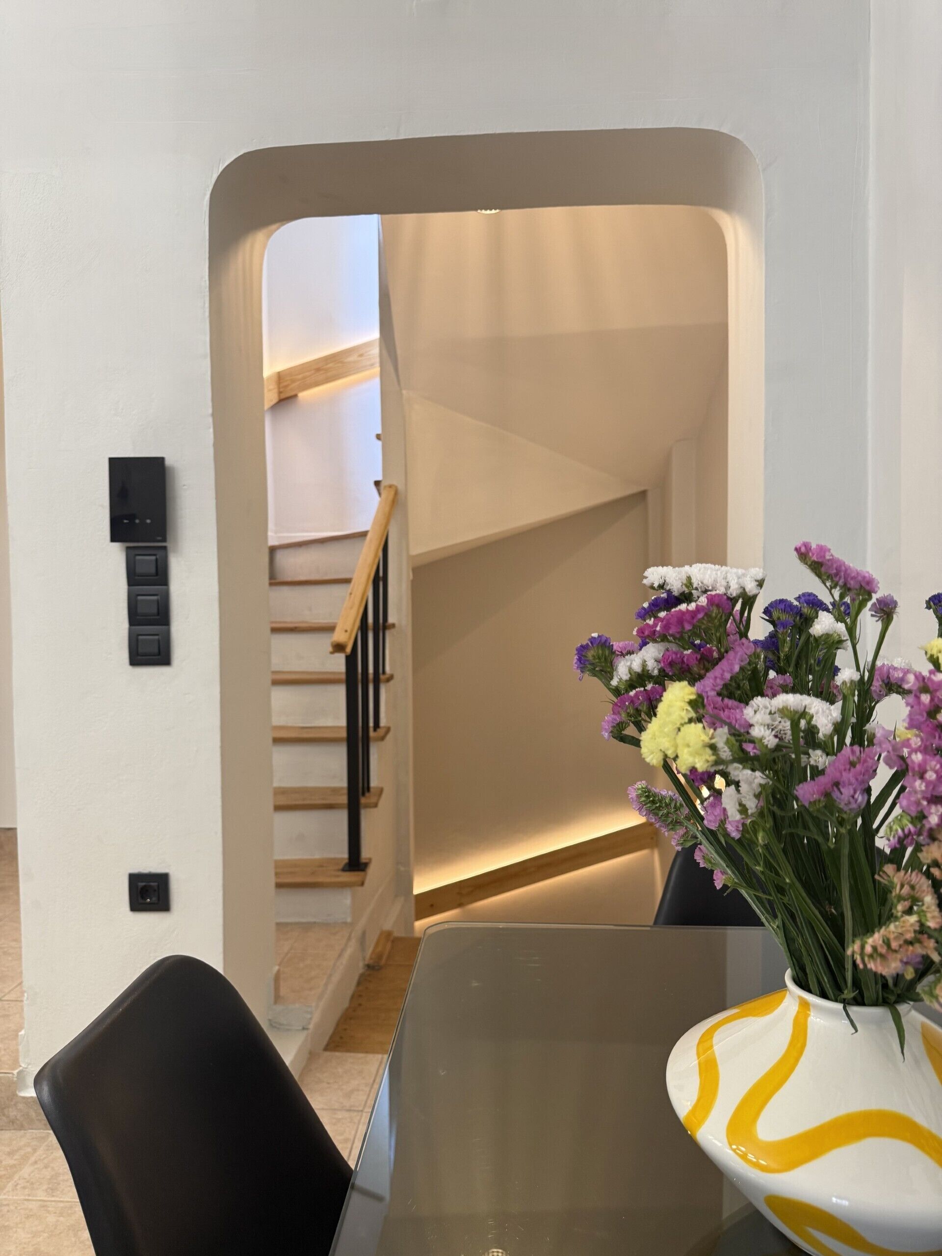Un interior moderno con una escalera, una zona de comedor con mesa de vidrio y un jarrón con flores.