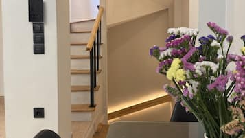 Un interior moderno con una escalera, una zona de comedor con mesa de vidrio y un jarrón con flores.