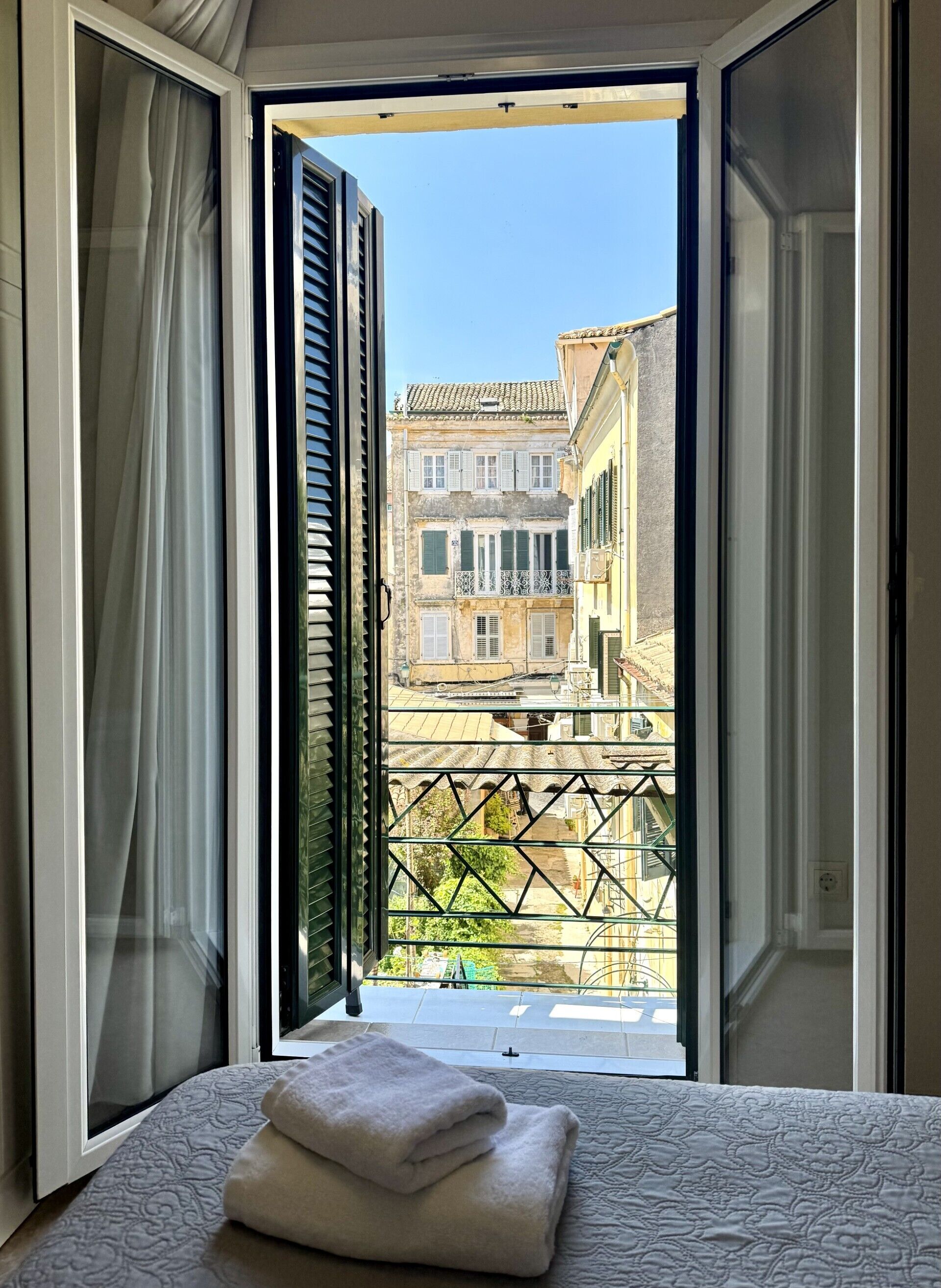 Una habitación con cama, ventana con contraventanas abiertas y vista a edificios.