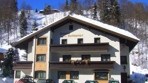 A multi-story building with a snow-covered roof, balconies, and parked cars in front.