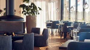 Een modern restaurantinterieur met ronde tafels, grijs gestoffeerde stoelen, een grote potplant en een houtkachel.