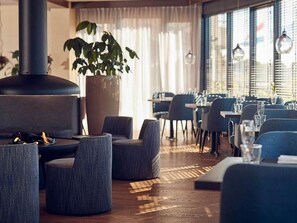A modern restaurant interior with round tables, grey upholstered chairs, a large potted plant, and a wood-burning stove. - 2 Comfy Rooms on Scenic Ameland – Near Beach, Ferry, Dunes & Cycling Routes (Nes)