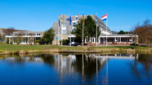 Een groot gebouw met meerdere vlaggen, een vijver ervoor en een helderblauwe lucht.