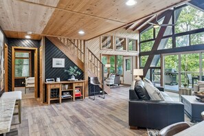 A spacious living area with a wooden staircase, a sofa, a dining table, and large windows offering a view of the outdoors. - Lakeside Getaway at Innsbrook by Sarah Bernard (Innsbrook)