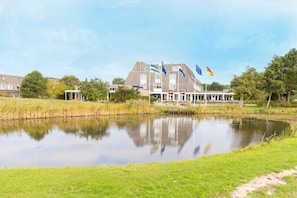 Un edifici amb diverses banderes, incloent-hi la bandera de la Unió Europea, un estany al davant i una zona enjardinada.
