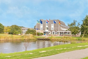A hotel with multiple flags, a pond, and a grassy area. - Stay on Ameland Near Beach – Hotel Comfort, Dining & Nature Walks - 4 Units (Nes)