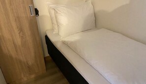 Una cama individual con ropa blanca en una habitación con puerta de madera y marco negro.