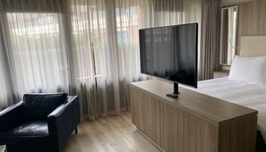 Et hotelværelse med en seng, et fjernsyn på et træskab, en sort læderstol og lette gardiner.